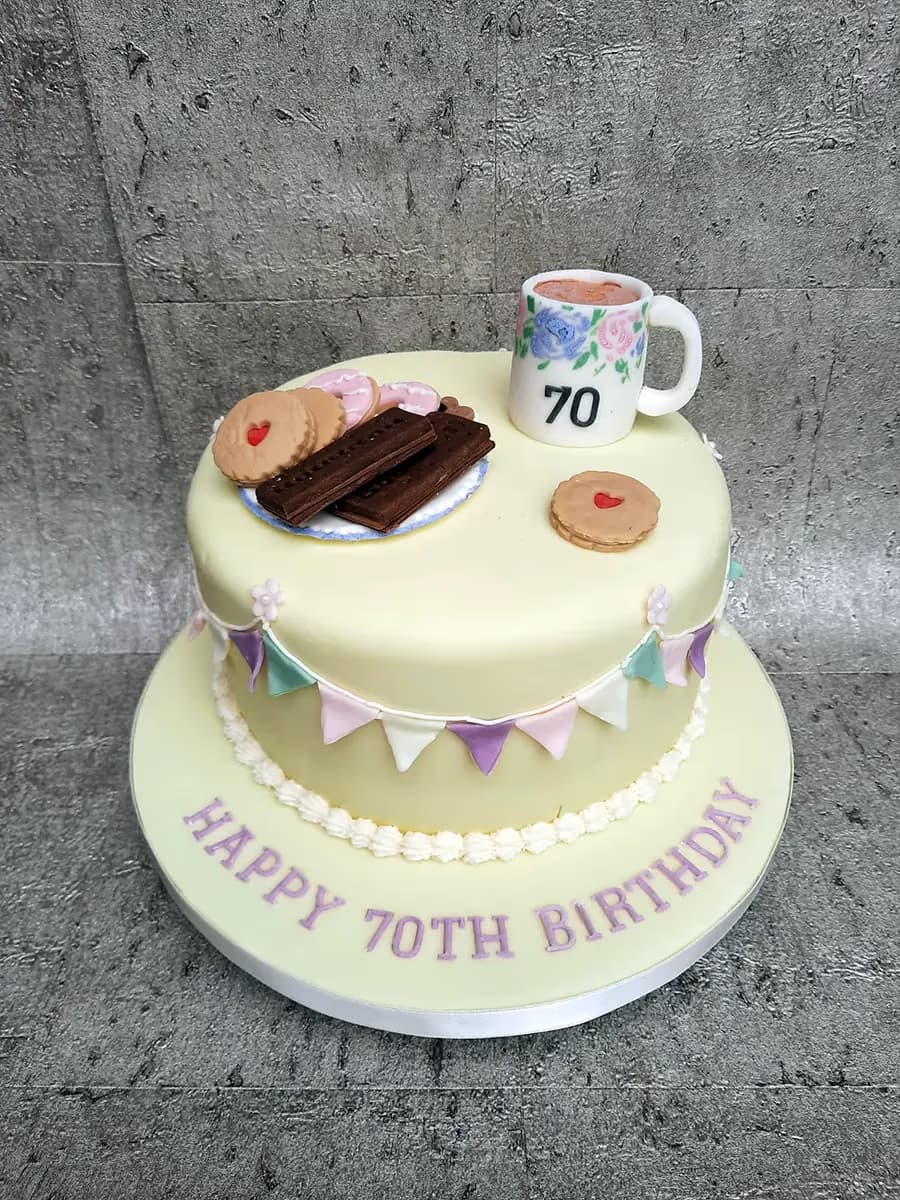 A two-tier pale yellow 70th birthday cake. The top tier is adorned with edible decorations including a small plate of biscuits (chocolate fingers, heart-shaped cookies, and pink iced ring biscuits), a miniature white mug with '70' and floral patterns, and a single heart-shaped cookie. Pastel bunting in mint green, pink, purple, and white wraps around the top tier. The base of the cake board reads 'HAPPY 70TH BIRTHDAY' in purple lettering, framed by a white piped border.