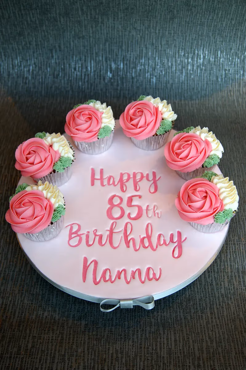 A light pink round cake board with seven cupcakes arranged in a semi-circle, each topped with a pink rose, white, and green frosting. The board has 'Happy 85th Birthday Nanna' written in a darker pink script, with a grey ribbon bow at the bottom.