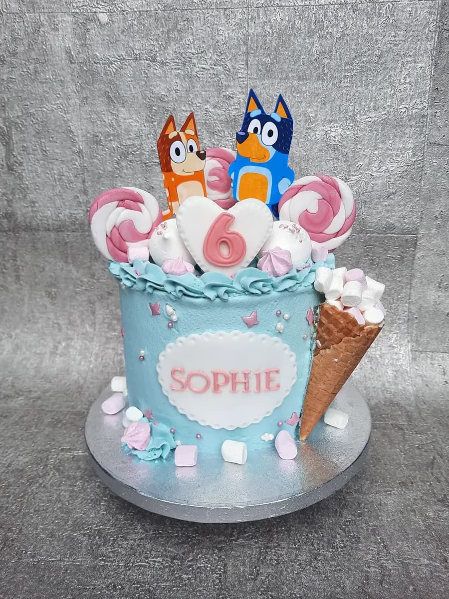 A light blue single-tier birthday cake featuring Bluey and Bingo character toppers, a white heart with a pink number 6, and the name 'SOPHIE' on the side. It is decorated with pink and white lollipops, meringues, marshmallows, and an ice cream cone filled with marshmallows.