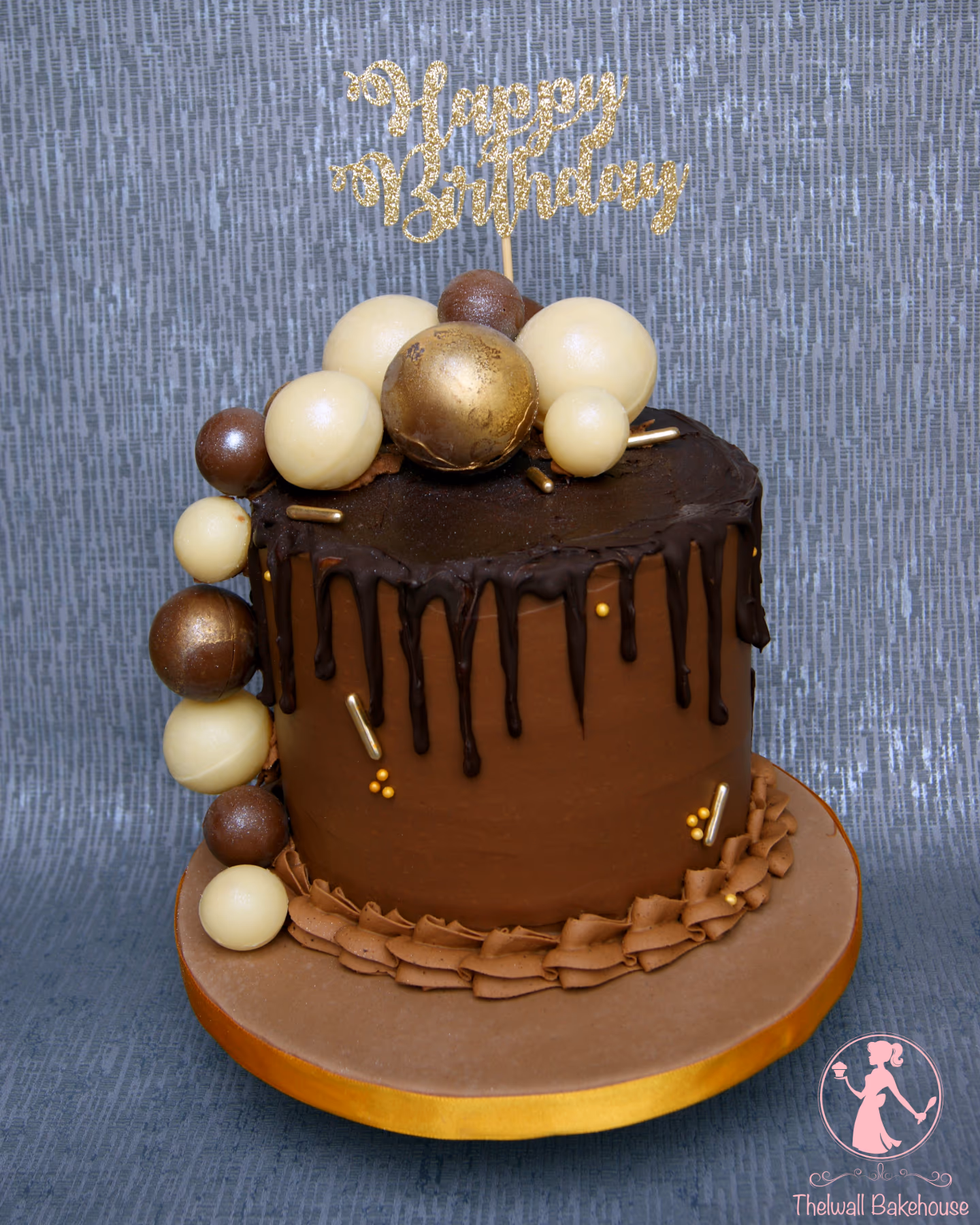 A tall, single-tier chocolate birthday cake with dark chocolate ganache drip, decorated with white, gold, and bronze chocolate spheres, gold sprinkles, and a gold glitter 'Happy Birthday' topper.