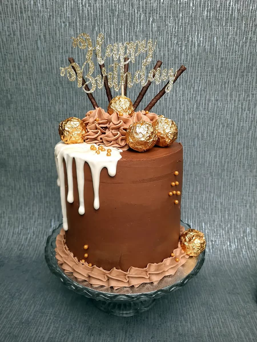 A single-tier chocolate birthday cake with white chocolate drip, topped with chocolate frosting swirls, Ferrero Rocher chocolates, gold sprinkles, and a gold glitter 'Happy Birthday' topper on a glass cake stand.