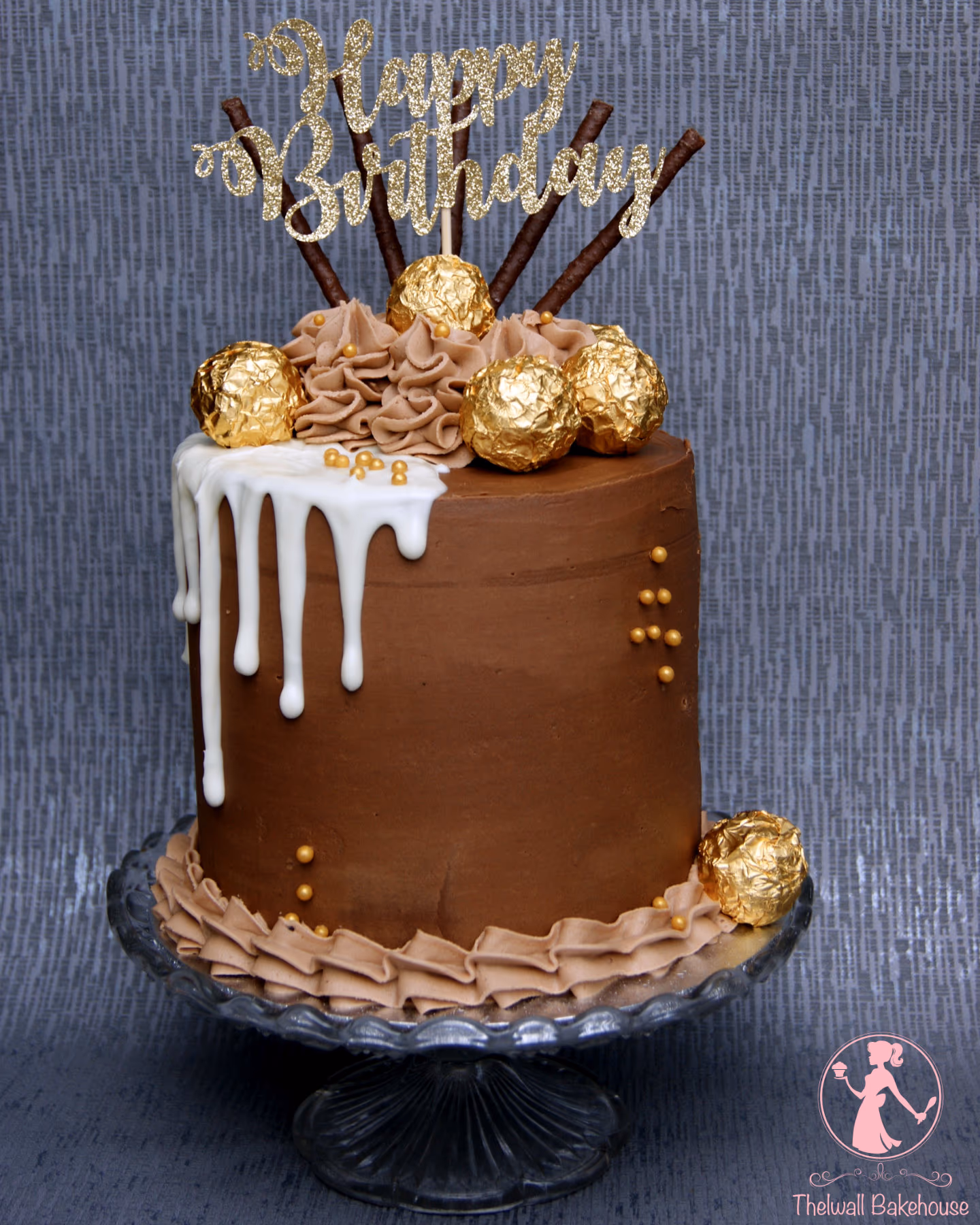 A chocolate birthday cake with white chocolate drip, decorated with gold-wrapped chocolates, chocolate wafer sticks, gold sprinkles, and a gold glitter 'Happy Birthday' topper, sitting on a clear glass cake stand.