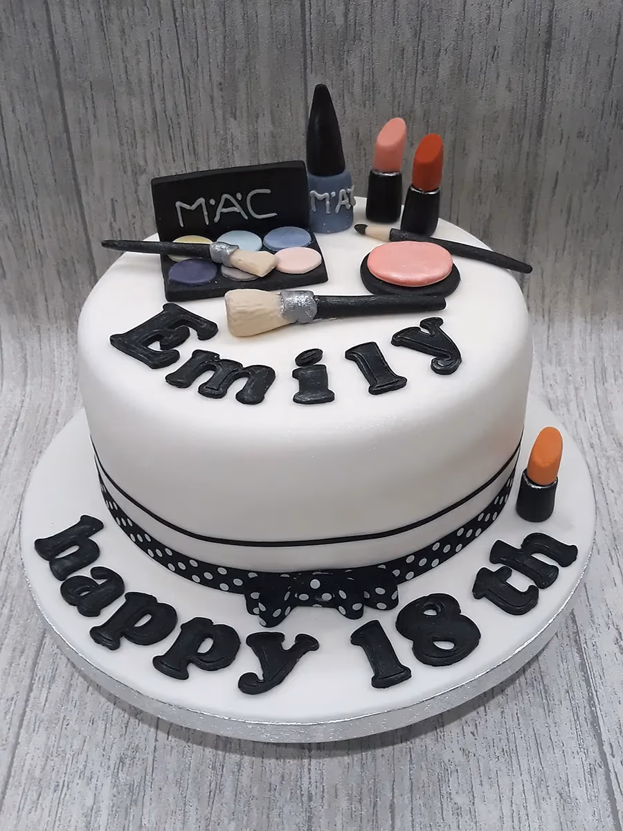 A white round birthday cake decorated with black fondant letters spelling 'Emily' on top, surrounded by makeup-themed decorations including a MAC eyeshadow palette, lipsticks, nail polish, and brushes. The base of the cake features black fondant letters spelling 'happy 18th' and a black and white polka dot ribbon with a bow.