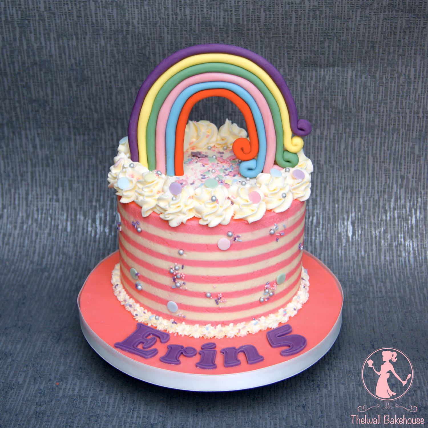 A single-tier round birthday cake for Erin's 5th birthday, decorated with pink and white horizontal stripes, a border of white buttercream swirls, colorful sprinkles, and a large rainbow made of fondant. The pink cake board has the name 'Erin 5' written in purple letters.
