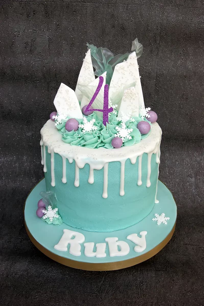 A Frozen-themed birthday cake for Ruby's 4th birthday, featuring a teal base, white drip icing, and topped with white chocolate shards, purple candies, and snowflakes, with a glittery purple number 4.