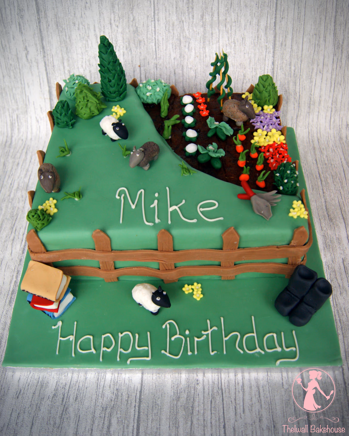 A square birthday cake decorated with an elaborate garden and farm scene. The top tier features a green hillside with trees, bushes, and sheep, next to a brown garden patch filled with various vegetables, colorful flowers, and a small rake. The name 'Mike' is written on the hill. The bottom tier includes a wooden fence, a stack of books, another sheep, and black boots, with 'Happy Birthday' written in white icing.