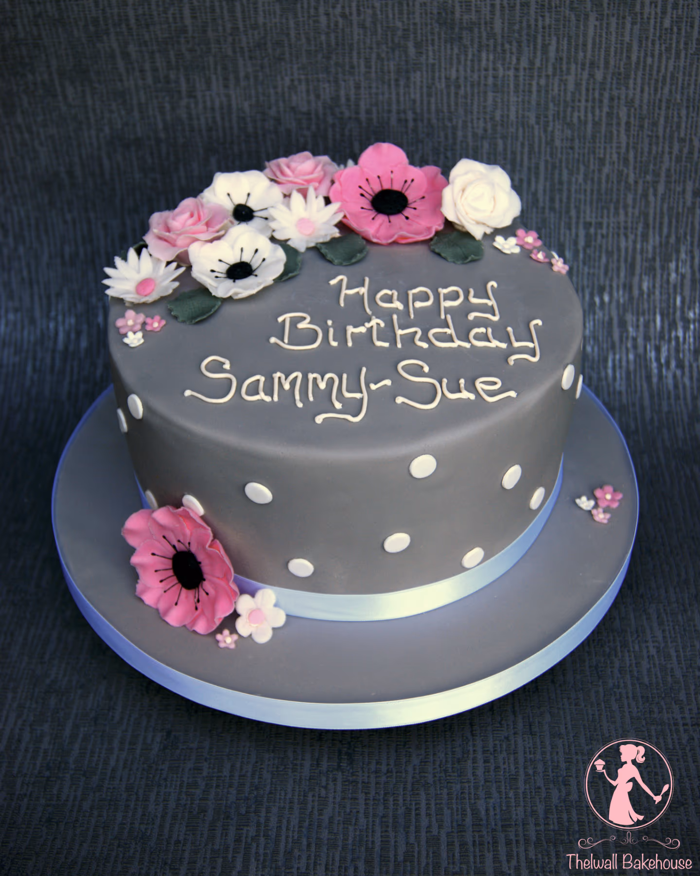 A round grey birthday cake decorated with pink and white sugar flowers, white polka dots, and a light blue ribbon, with 'Happy Birthday Sammy-Sue' written in white icing.
