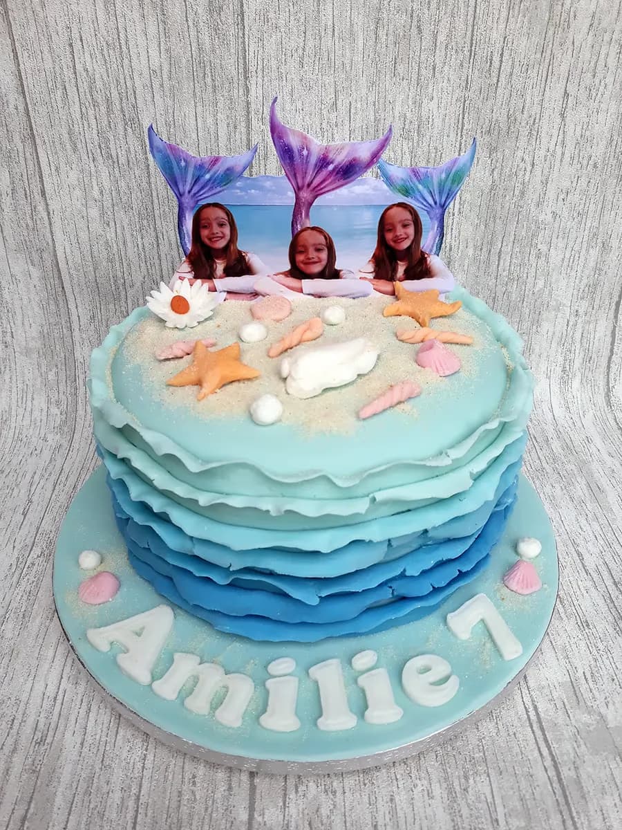 A two-tier mermaid-themed birthday cake featuring blue ombre ruffles on the bottom tier, edible sand, seashells, and orange starfish on the top tier, and three photo toppers of girls with mermaid tails. The light blue cake board reads 'Amilie 7' in white letters.