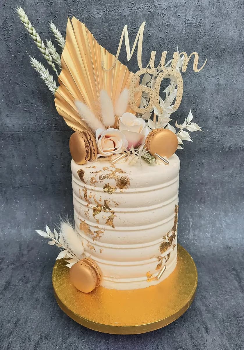 A single-tier cream birthday cake with ribbed frosting and gold leaf, topped with a gold 'Mum 60' topper, a gold fan, dried white wheat, fluffy accents, pale pink roses, and gold macarons, all on a gold cake board.