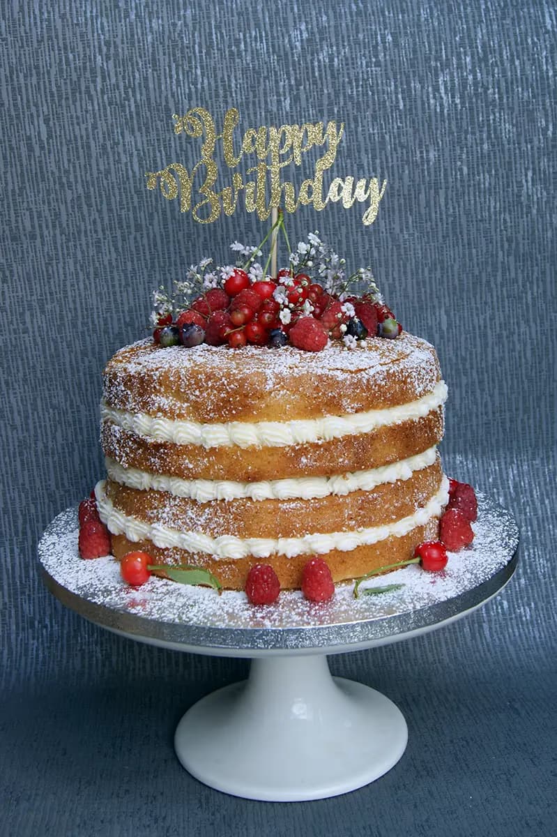 A three-tier naked birthday cake with white cream filling between sponge layers, topped with fresh mixed berries, small white flowers, and a gold glitter 'Happy Birthday' cake topper, dusted with powdered sugar and presented on a white pedestal stand.