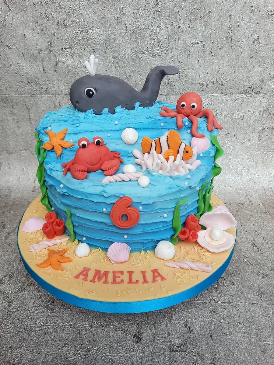 An ocean-themed birthday cake with blue wave-like icing, decorated with fondant sea creatures including a whale, red crab, orange octopus, and clownfish, along with starfish, shells, the number 6, and the name Amelia on the sandy base.