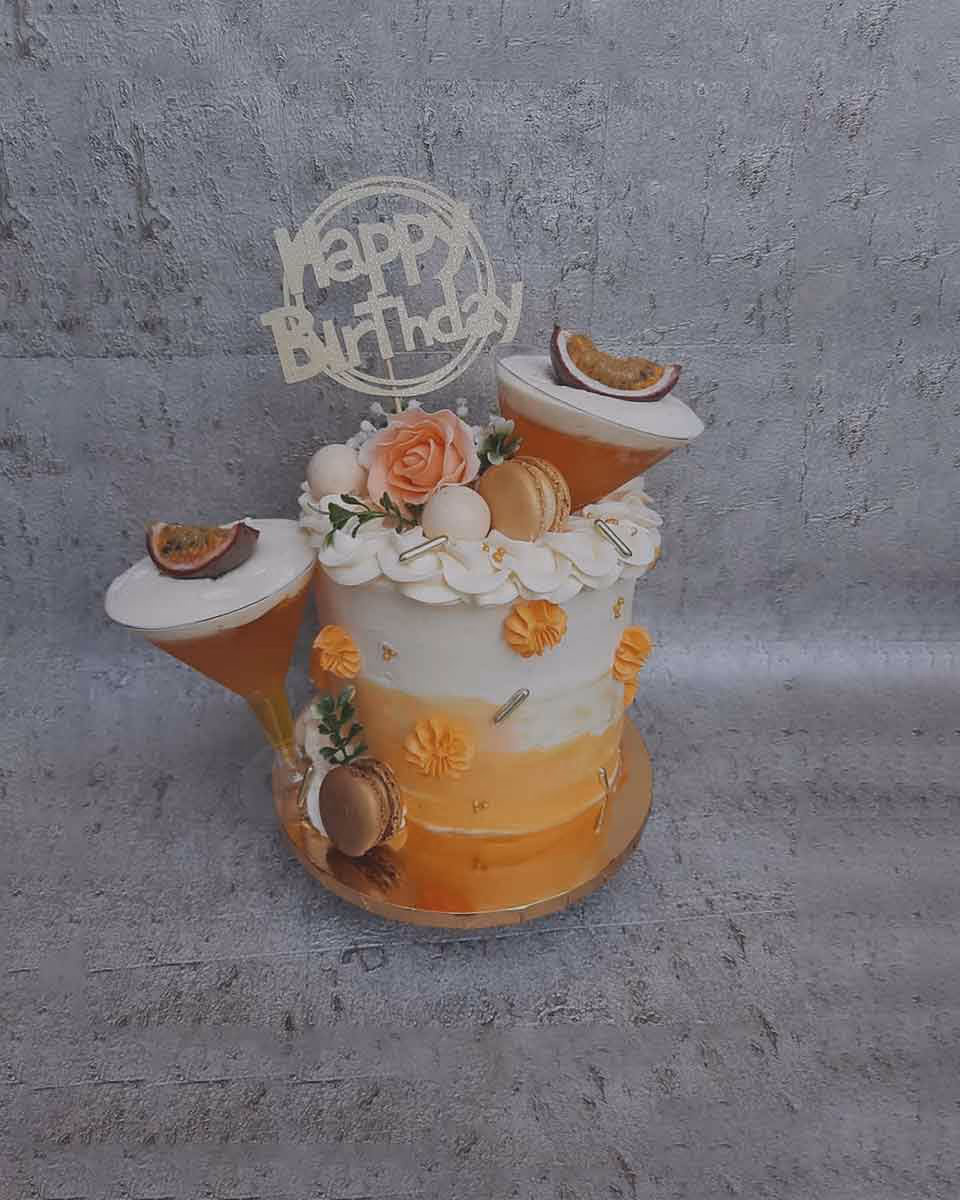 A single-tier ombré birthday cake, transitioning from white to orange, is topped with a 'Happy Birthday' topper. It's decorated with white buttercream swirls, an orange sugar rose, white spheres, macarons, and two tilted martini glass-shaped elements, each holding a passion fruit slice and appearing to pour orange liquid. Small orange sugar flowers and silver sprinkles decorate the sides.