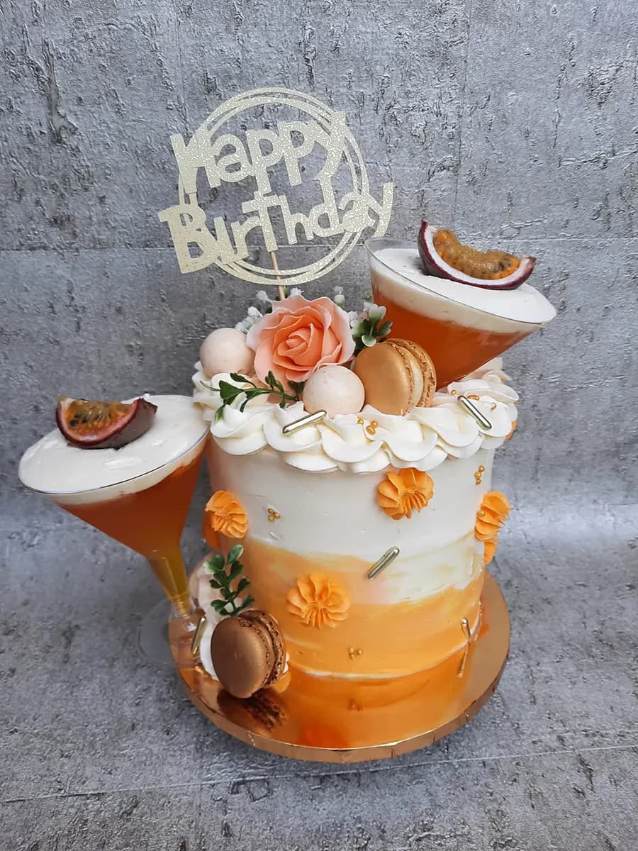 A single-tier birthday cake featuring an orange ombre design fading into white buttercream. It's decorated with white frosting swirls, an orange sugar rose, gold macarons, small white spheres, and orange rosettes. Two tilted clear plastic martini glasses, filled with an orange drink, white cream, and a passion fruit slice, are placed on and next to the cake. A gold 'Happy Birthday' topper sits at the top.