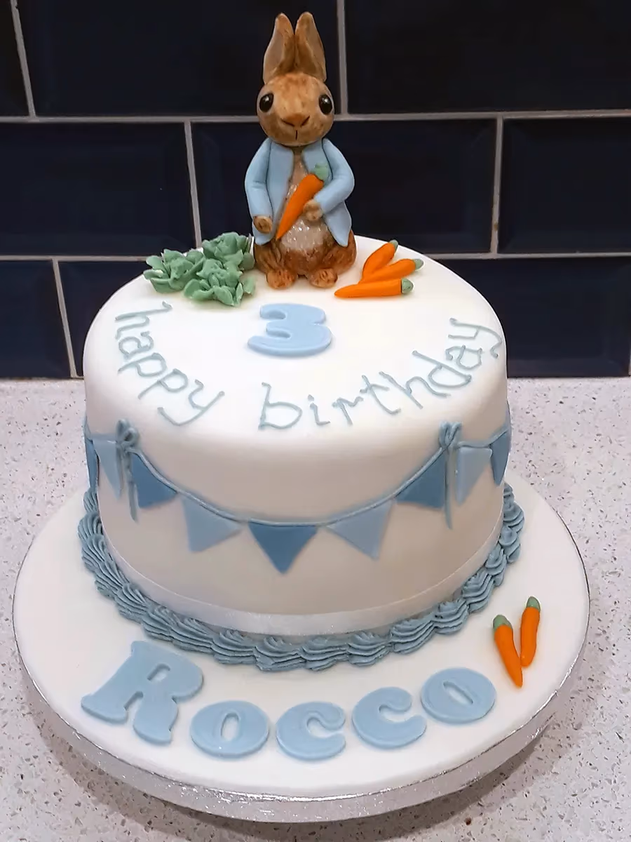 A white round birthday cake featuring a Peter Rabbit figurine holding a carrot, a light blue fondant number '3', 'happy birthday' text, a blue bunting banner, and the name 'Rocco' in blue fondant letters on the cake board, with additional fondant carrots and green foliage.