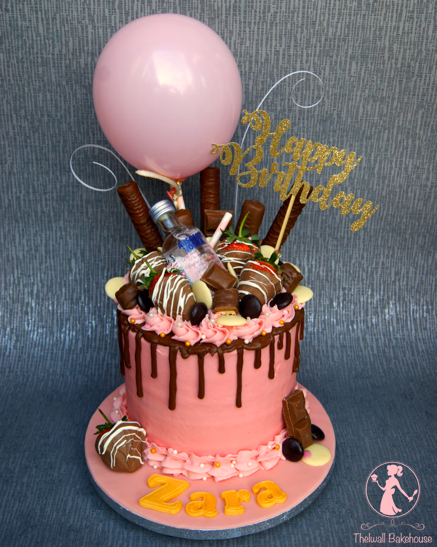 A pink birthday drip cake with dark chocolate ganache, topped with a mini Absolut Vodka bottle, chocolate-dipped strawberries, various chocolates, pink buttercream, a pink balloon, and a gold glitter 'Happy Birthday' topper. Gold letters spelling 'Zara' are on the pink cake board.