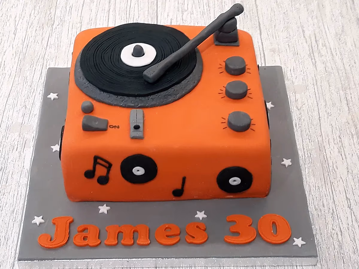 An orange record player shaped birthday cake with grey details, featuring a black vinyl and tonearm, decorated with musical notes and white stars, sitting on a grey board with "James 30" written in orange lettering.