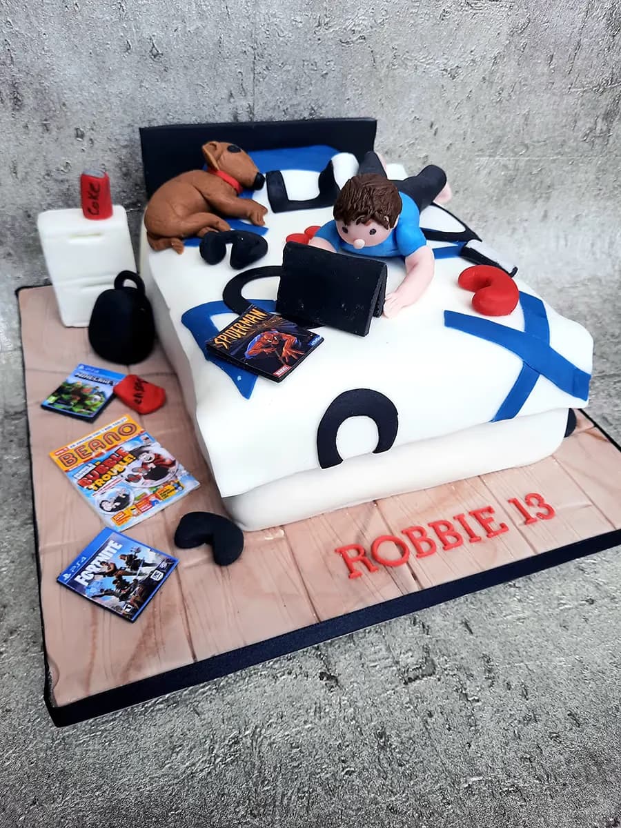 A rectangular birthday cake designed as a teenager's bedroom, featuring a boy on a bed with a laptop, a sleeping brown dog, a bedside table with a Coke can, and various items like Spiderman, Minecraft, Fortnite, and Beano comics on the wooden-effect base. The name "ROBBIE 13" is written in red letters on the base.