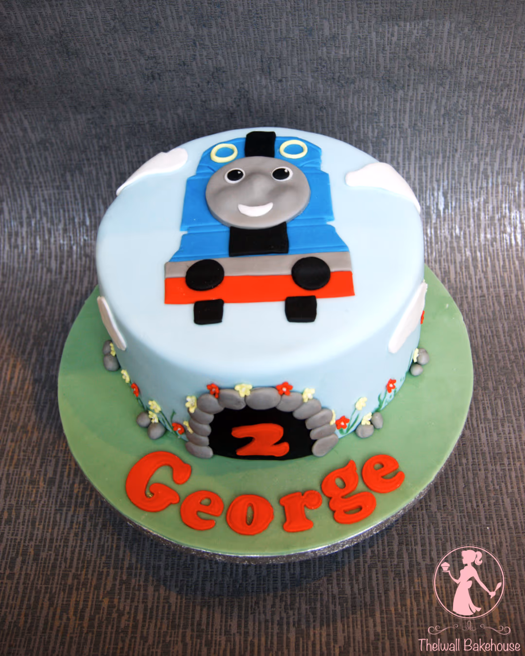 A light blue round birthday cake decorated with Thomas the Tank Engine on top, white clouds, and a green base. The side of the cake features a grey stone tunnel with the number '2' in red, surrounded by small red and yellow flowers and the name 'George' spelled out in red fondant letters on the green base.