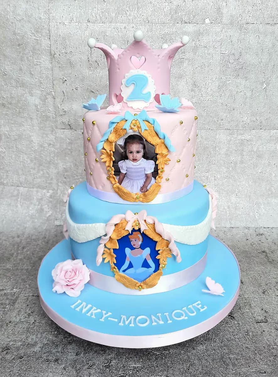 A three-tier birthday cake in pink and light blue. The top tier features a pink crown with the number 2. The middle tier has a quilted design and a framed picture of a young girl. The bottom tier includes a framed image of Cinderella. The light blue base is adorned with a pink ribbon, a pink rose, a butterfly, and the name 'INKY-MONIQUE' in white letters.