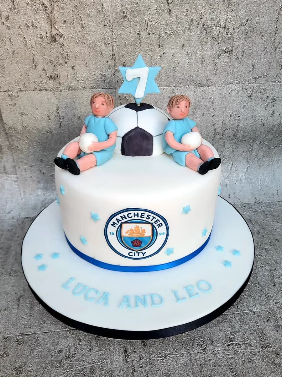 A two-tier white birthday cake for twins, decorated with two boy figurines in light blue soccer jerseys, a soccer ball, a light blue star with the number 7, the Manchester City logo, and the names 'Luca and Leo' on the base.