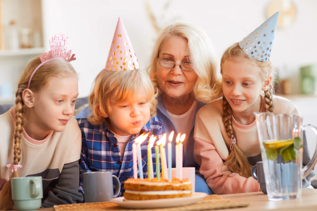 A young boy in a party hat blows out the candles on a two-layer birthday cake, watched by his grandmother and two sisters.