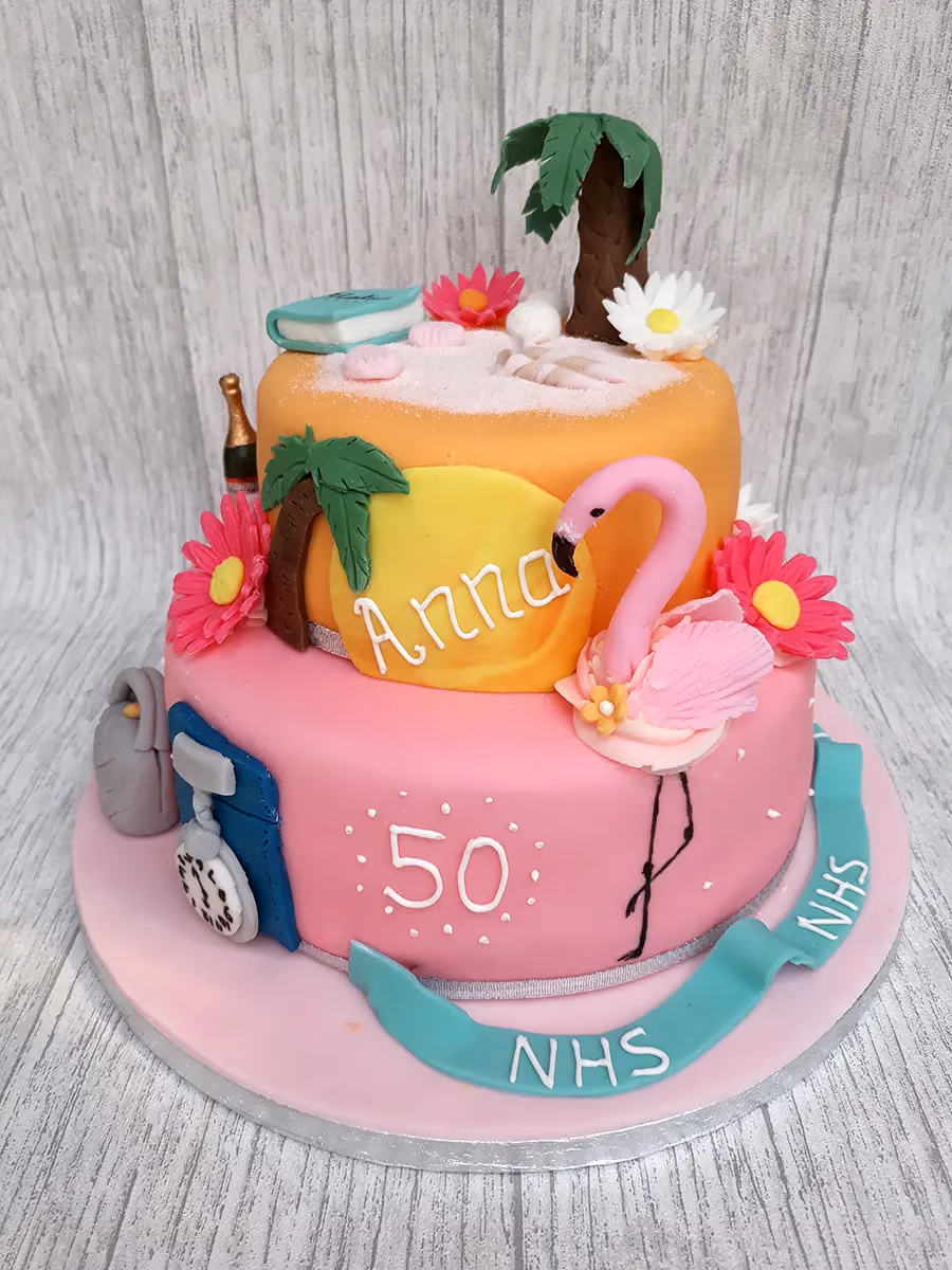 A two-tier birthday cake. The top tier is orange and decorated as a sandy beach with palm trees, a book, seashells, and a champagne bottle. The bottom pink tier features a flamingo, a yellow plaque with 'Anna' written on it, the number '50', and nursing-themed decorations like a stethoscope and a uniform, with a teal ribbon displaying 'NHS'.