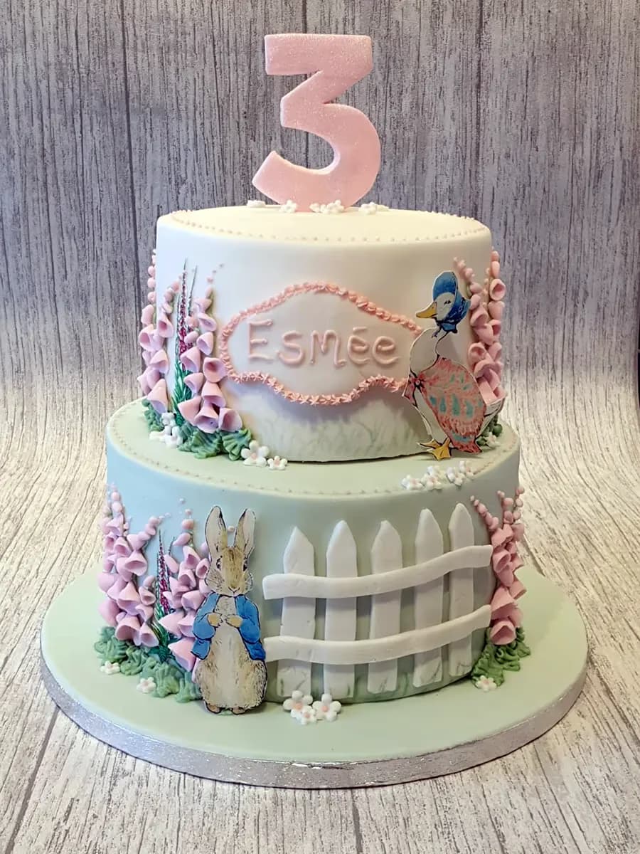 A two-tier Peter Rabbit themed birthday cake with white and pale green fondant, featuring a pink glittery number three on top, the name Esmée, illustrations of Peter Rabbit and Jemima Puddle-Duck, pink foxglove flowers, and a white picket fence.