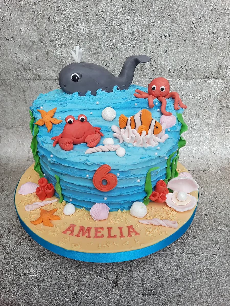 An under-the-sea themed birthday cake with blue rippled icing, featuring a gray whale, red crab, orange octopus, and clownfish, adorned with edible coral, starfish, seashells, and pearls, with a red number '6' and the name 'AMELIA' on the sandy base.