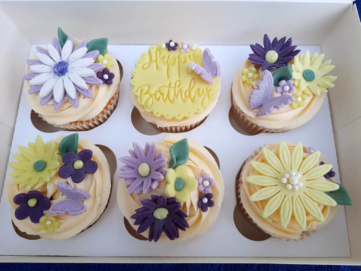 Six pastel-colored cupcakes in a white box, featuring cream frosting and decorated with sugar flowers and butterflies in shades of purple, lavender, and yellow. One cupcake has a yellow round plaque with 'Happy Birthday' written on it.