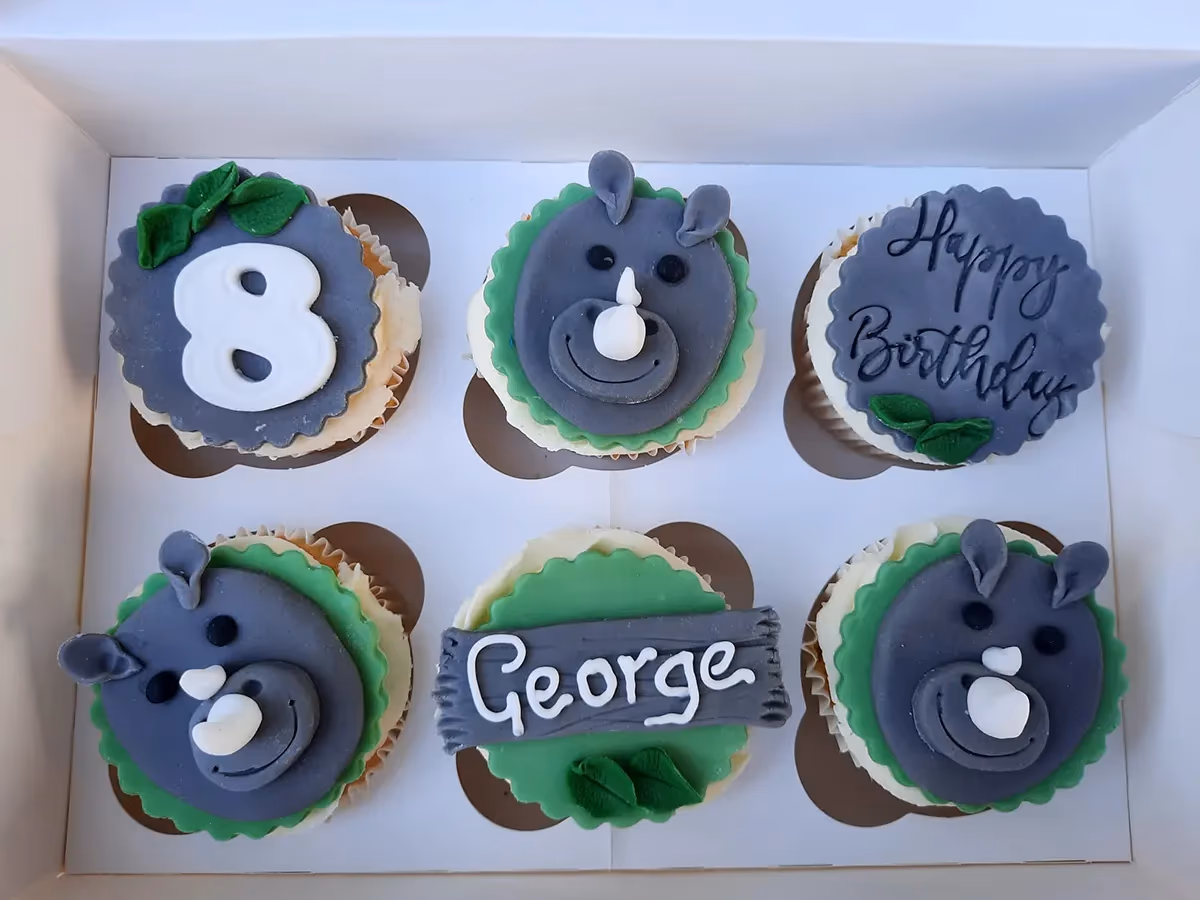 Six birthday cupcakes in a white box, featuring three grey rhino faces, one cupcake with 'Happy Birthday' text, one with the number '8', and one with 'George' text, all with green and grey fondant decorations.
