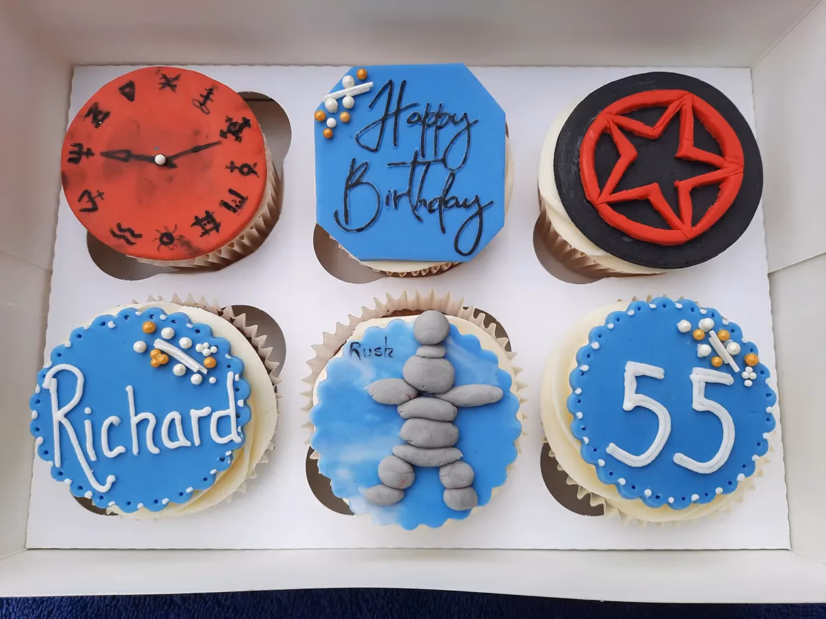 A white box containing six themed birthday cupcakes. The top row features a red clock face with black symbols, a blue octagonal cupcake with 'Happy Birthday' in black script, and a black cupcake with a red five-pointed star. The bottom row shows a blue scalloped cupcake with 'Richard' in white script, a light blue cupcake with a grey Inukshuk figure and 'Rush' written on it, and a blue scalloped cupcake with '55' in white numbers.