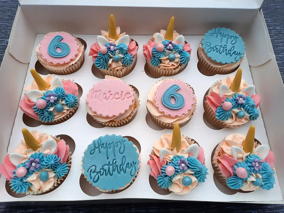A box of twelve pastel-colored birthday cupcakes, some decorated as unicorns with horns and floral frosting, and others with round fondant toppers displaying '6', 'Marcie', and 'Happy Birthday'.
