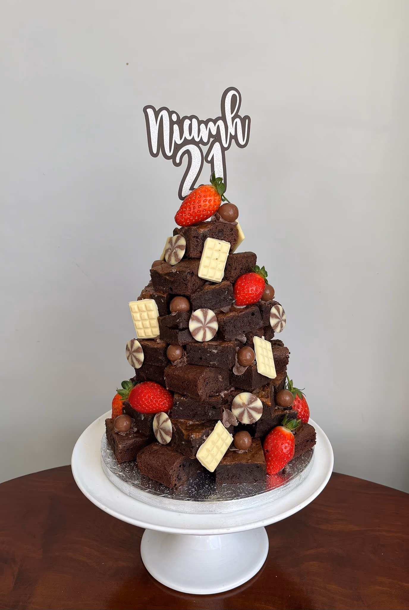 A pyramid-shaped brownie stack cake, decorated with fresh strawberries, white chocolate bars, chocolate swirl discs, and chocolate balls, topped with a 'Niamh 21' cake topper, presented on a white cake stand.