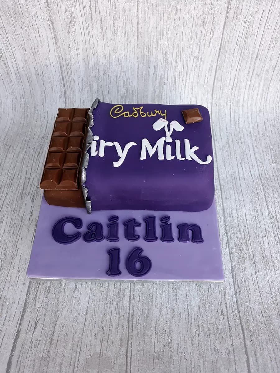 A rectangular birthday cake shaped like a Cadbury Dairy Milk chocolate bar, covered in purple fondant with 'Cadbury Dairy Milk' written in white and gold. A section of brown chocolate is visible on the left side of the cake, as if the wrapper is peeled back. Below the cake, on a lighter purple board, are the words 'Caitlin' and the number '16' in purple.