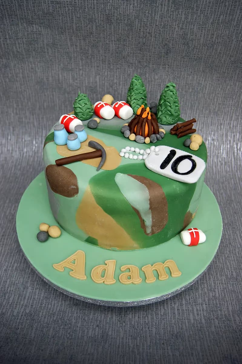 A round birthday cake with a green, brown, and light green camouflage pattern, decorated with edible camping-themed elements including a campfire, green pine trees, blue canteens, a pickaxe, and a white dog tag with the number '10'. The cake sits on a light green board with the name 'Adam' spelled in gold letters.