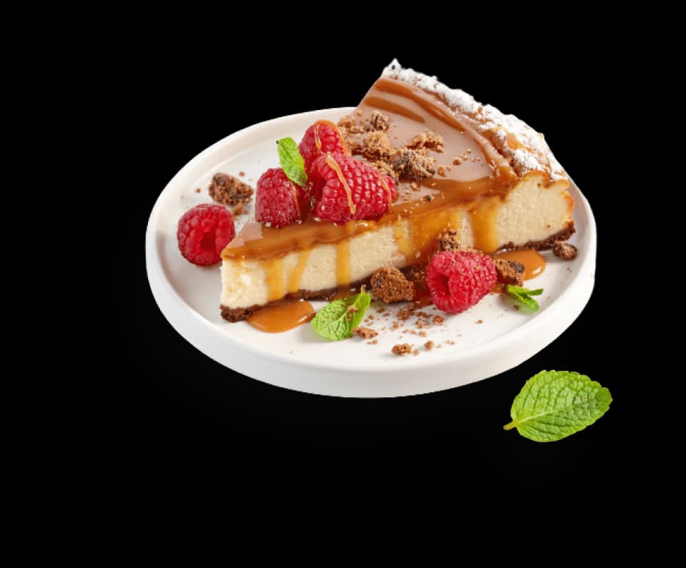 A slice of cheesecake topped with rich caramel sauce, fresh red raspberries, chocolate cookie crumbs, and garnished with mint leaves, served on a white plate.