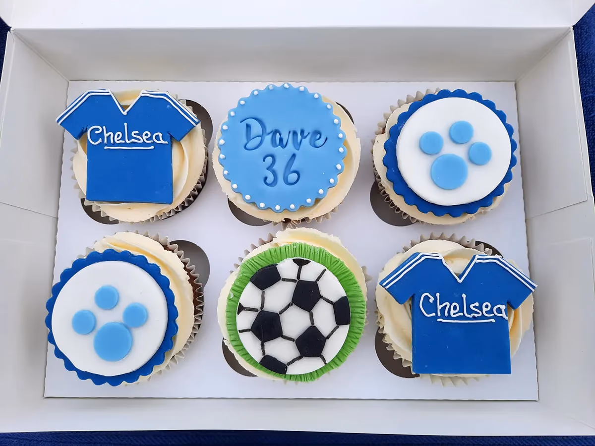 Six Chelsea FC themed cupcakes in a white box, featuring blue football shirts, a football, blue paw prints, and one with 'Dave 36' written in white icing.