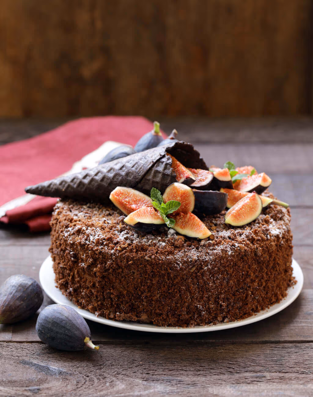 A round chocolate crumb cake topped with powdered sugar, sliced fresh figs, whole figs, a dark chocolate waffle cone, and fresh mint leaves, set on a white plate on a wooden table.