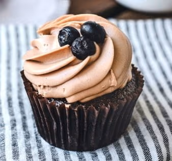 A dark chocolate cupcake with swirled chocolate frosting, topped with three fresh blueberries, sitting on a striped linen cloth.