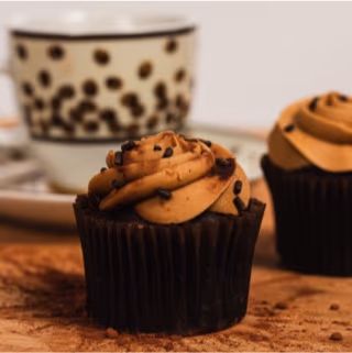 A close-up of a chocolate cupcake topped with swirled coffee-flavored frosting and chocolate sprinkles, with a blurred coffee-bean patterned cup and another cupcake in the background.