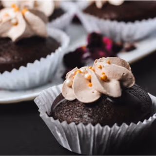 Close-up of a chocolate cupcake with swirled tan frosting and gold sprinkles in a white paper liner, with more cupcakes blurred in the background on a white serving tray.
