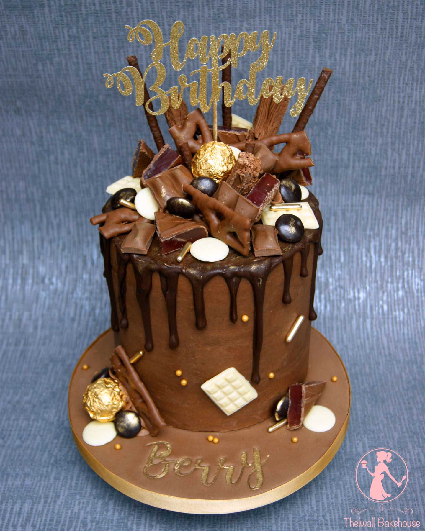 A rich chocolate drip birthday cake topped with an array of chocolate bars, truffles, and white chocolate discs, featuring a gold glittery 'Happy Birthday' topper and the name 'Berry' in gold on the base.