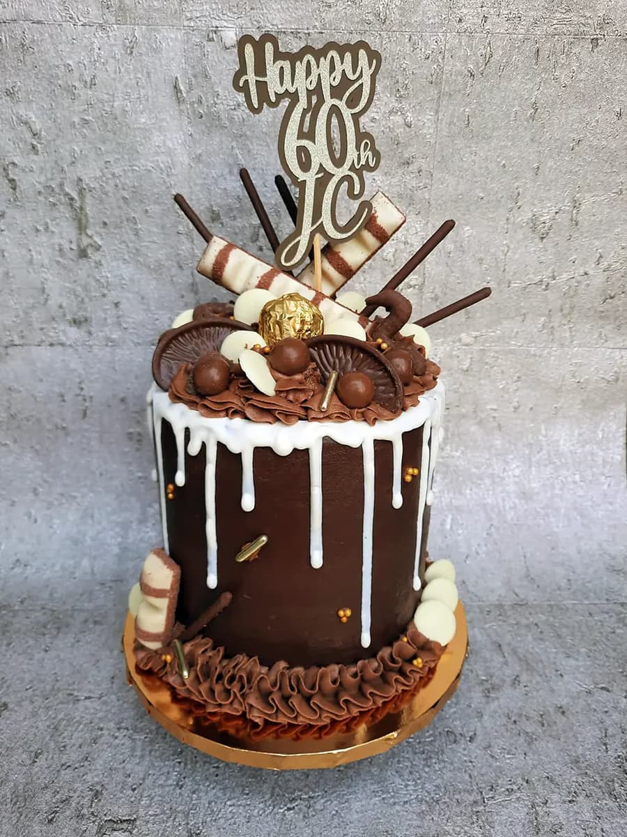 A rich dark chocolate drip cake with white chocolate ganache drips, decorated on top with various chocolates including chocolate balls, chocolate mushrooms, a gold Ferrero Rocher, and Kinder Bueno bars, and featuring a 'Happy 60th JC' birthday cake topper. The base of the cake has a piped chocolate buttercream border.