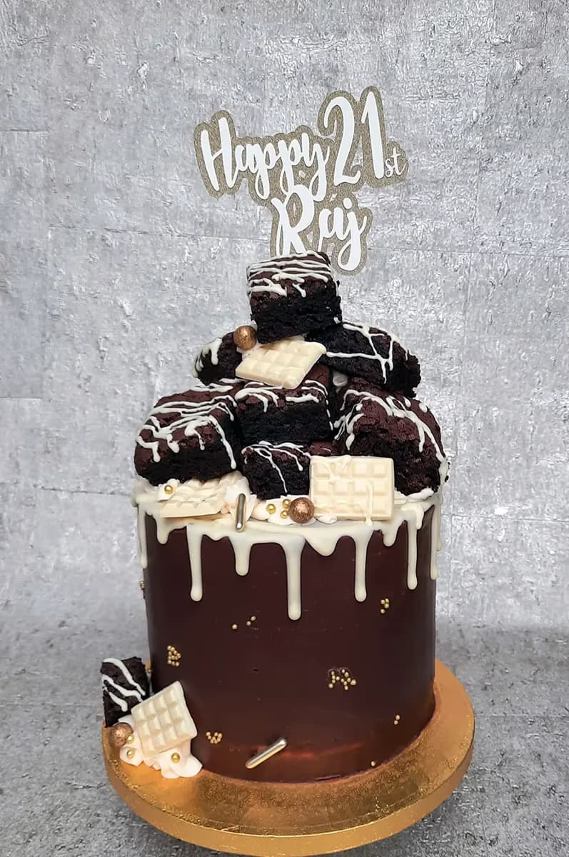 A single-tier dark chocolate cake with white drip icing, topped with a mound of brownies, white chocolate pieces, gold sprinkles, and a 'Happy 21st Raj' cake topper.