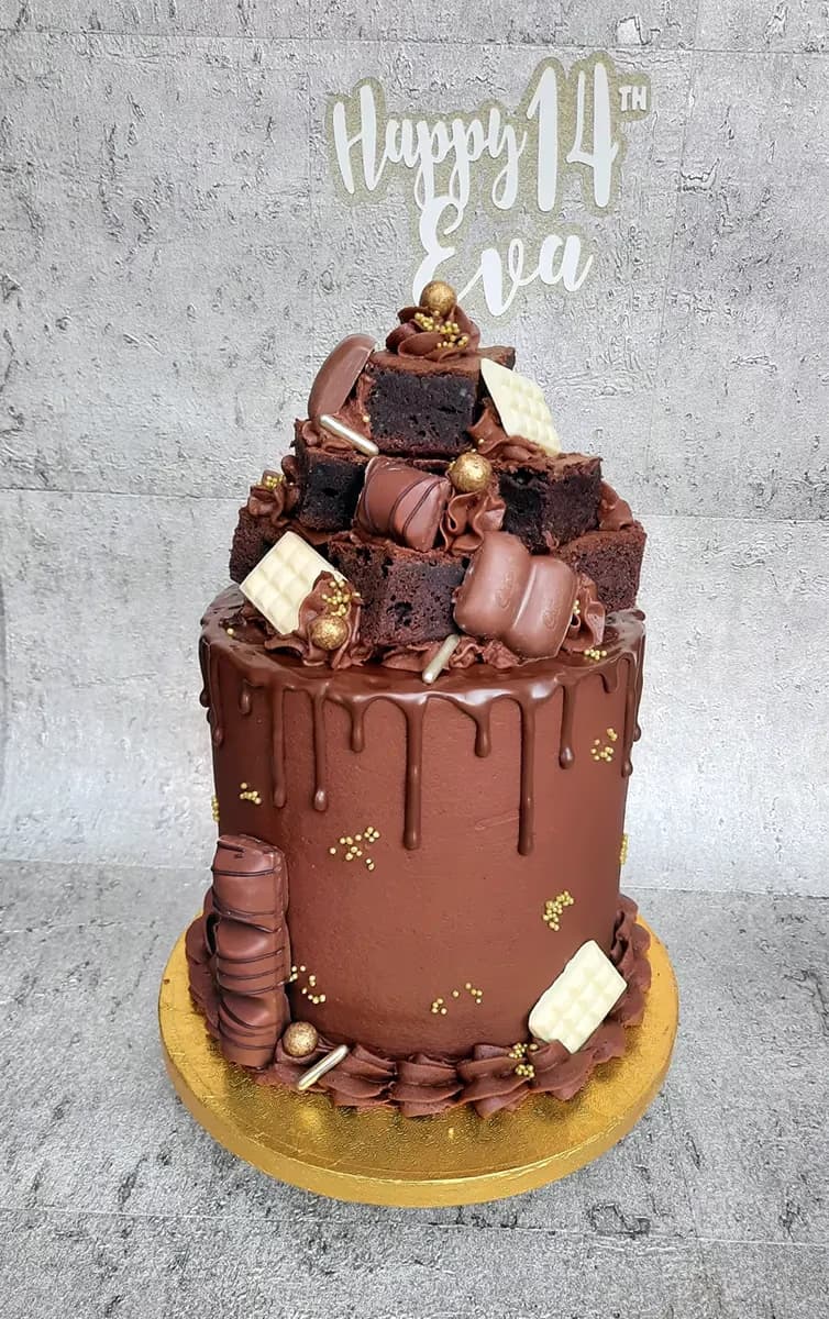 A tall, single-tier chocolate drip cake covered in a cascade of chocolate brownies, various milk and white chocolates, and gold sprinkles, with a 'Happy 14th Eva' cake topper at the top.