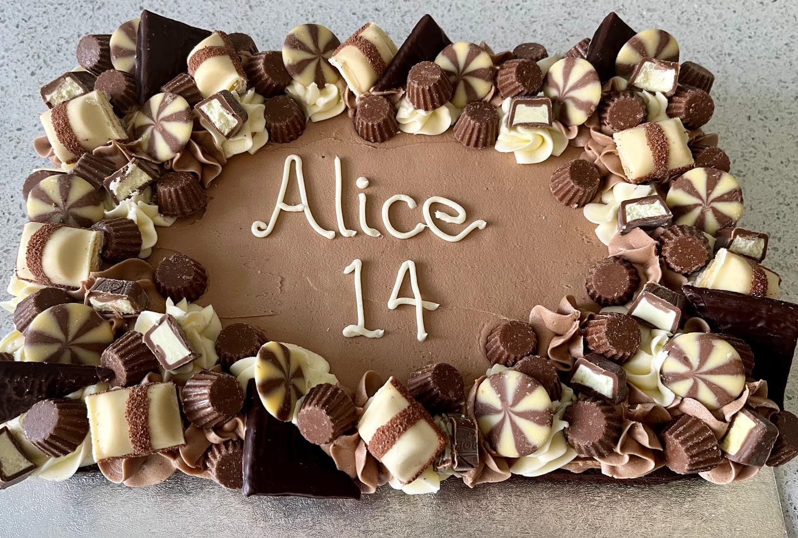 A rectangular chocolate birthday cake with 'Alice 14' written in white icing, decorated with a border of assorted chocolates including Kinder bars, chocolate cups, and swirl candies, interspersed with piped white and brown frosting.