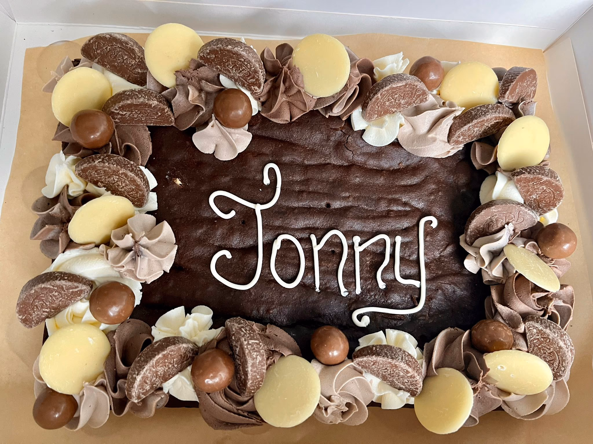 A rectangular chocolate brownie or cake, decorated with a border of white and milk chocolate frosting swirls, topped with milk chocolate segments, white chocolate buttons, and chocolate malt balls, with the name 'Jonny' written in white icing in the center.