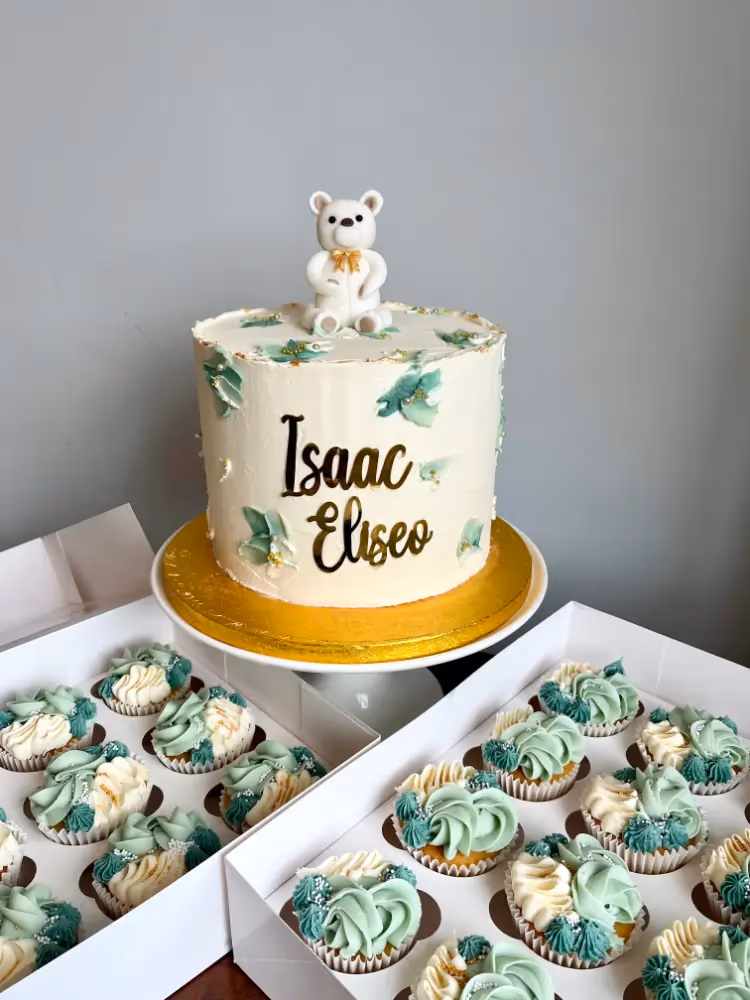 A white buttercream cake decorated with green leaf accents and a white teddy bear topper, featuring the names 'Isaac Eliseo' in gold script, displayed with two boxes of matching green and white frosted cupcakes.