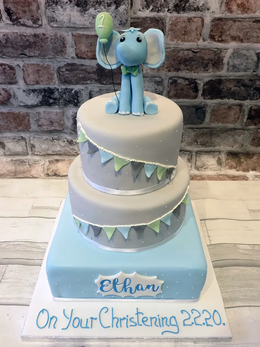 A two-tier christening cake in blue and grey, topped with a blue elephant holding a green balloon. The cake is decorated with blue and green bunting, white polka dots, and features the name 'Ethan' on the bottom tier. The cake board reads 'On Your Christening 2.2.20.'