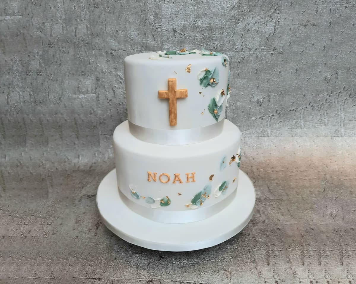 A two-tier white christening cake featuring a gold cross on the top tier and the name 'NOAH' in gold on the bottom tier, both decorated with abstract green, white, and gold accents, and white ribbons.